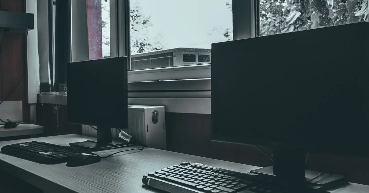 Espace de travail de bureau vide avec deux écrans d'ordinateur près d'une fenêtre, exprimant un environnement moderne et professionnel.