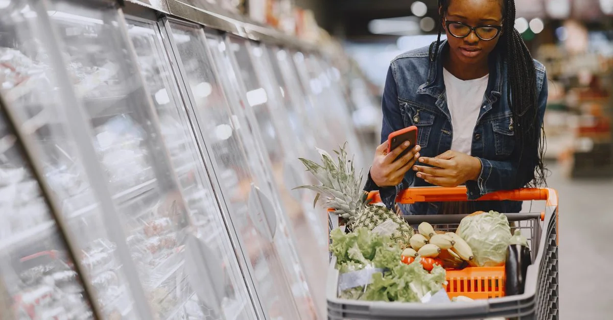 Femme dans un supermarché utilisant un smartphone tout en poussant un chariot de courses rempli de provisions.
