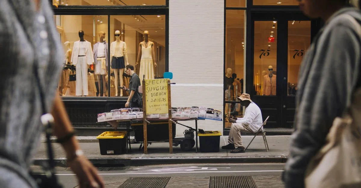 Un vendeur ambulant vendant des lunettes de soleil devant une vitrine de mode avec des mannequins.