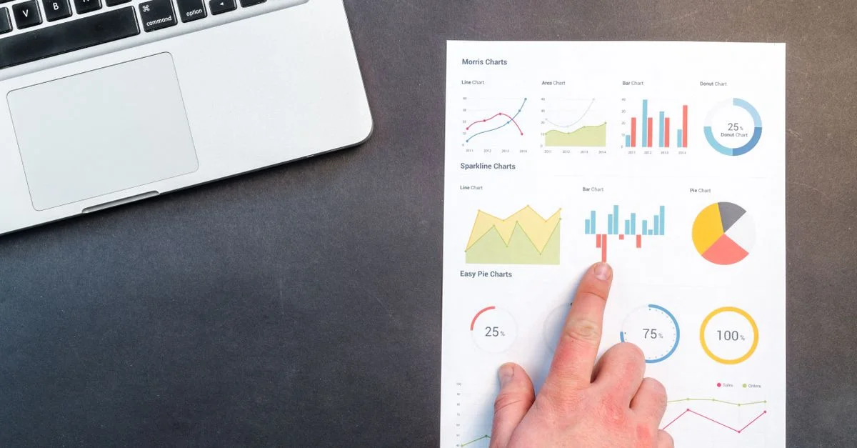 Une main pointant vers des graphiques colorés avec un ordinateur portable en arrière-plan sur un bureau.