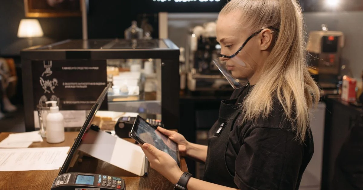Une barista portant un écran facial utilise une tablette pour travailler au comptoir d'un café, mettant en avant le service moderne.